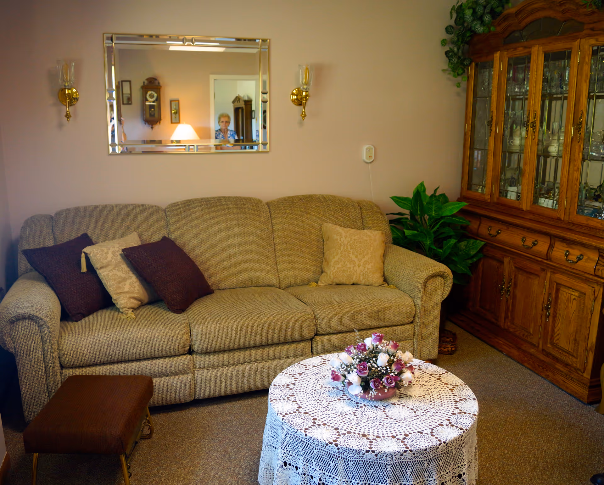 A cozy living room with a beige patterned sofa adorned with four pillows, a small brown footstool, and a round table covered with a white lace tablecloth and a floral centerpiece. On the right side, there is a wooden cabinet with glass doors displaying dishes and glassware. A large mirror hangs on the wall above the sofa, reflecting a lamp, a clock, and a person standing in the background. The walls are light-colored, and there are two wall-mounted candle holders on either side of the mirror.