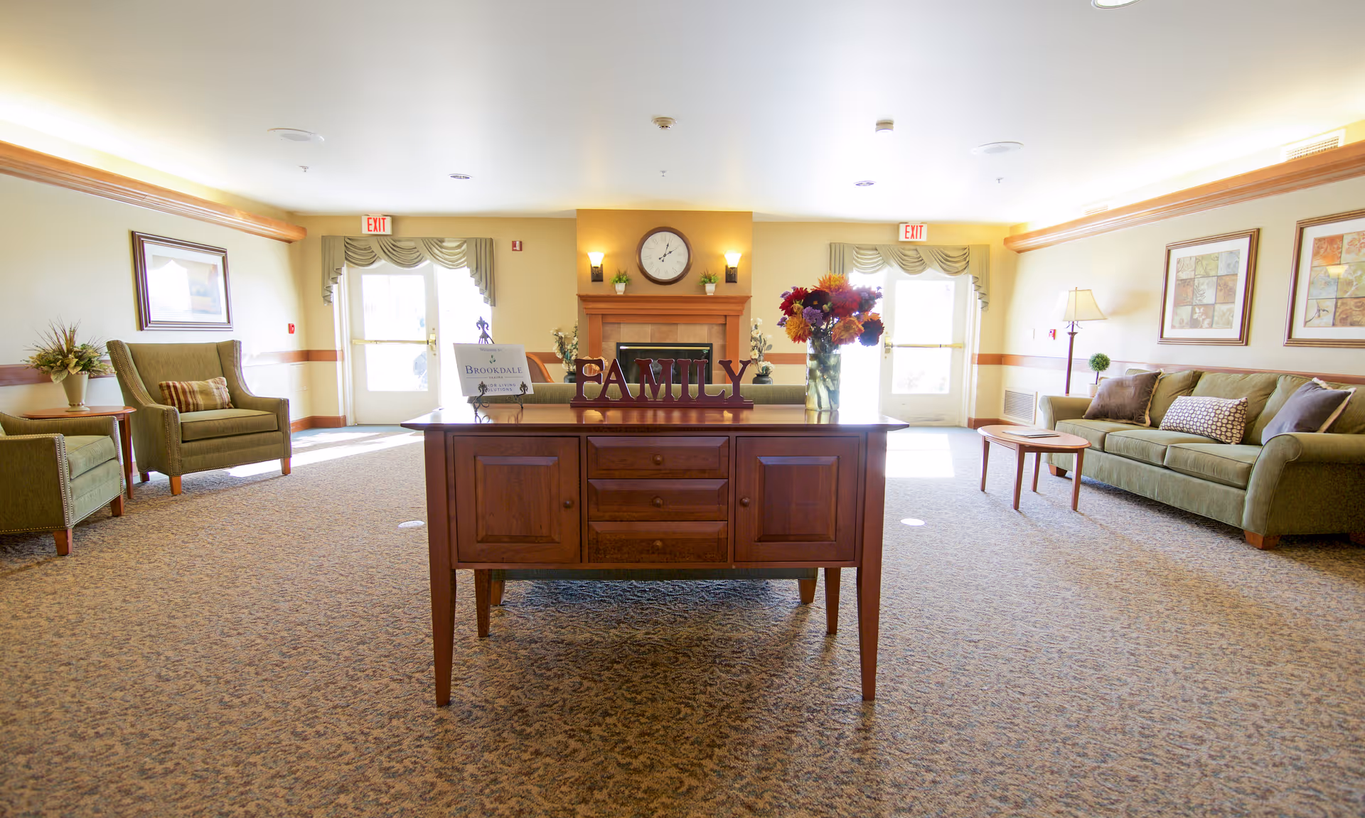 A bright and spacious living room area in a senior living facility with a wooden table in the center displaying a decorative 'FAMILY' sign and a vase of flowers. The room features a fireplace with a clock above it, two exit doors with windows, green upholstered chairs and a sofa, framed artwork on the walls, and a carpeted floor.
