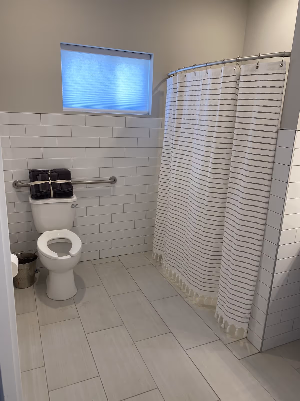 A clean and modern bathroom with light gray tiled floor and white subway tiled walls. There is a toilet with a raised seat and a grab bar behind it, with a stack of dark towels placed on top of the toilet tank. To the right, there is a shower area with a white shower curtain featuring horizontal gray stripes. A frosted window is above the toilet, allowing natural light into the room.