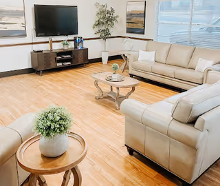 A bright communal living room with beige leather sofas, round coffee tables, a TV on a media console, and wood flooring.