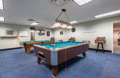 Interior view of a recreational room with two pool tables, overhead lights, a dartboard on the wall, and several chairs. The room has blue carpet flooring and light-colored walls with some framed pictures and a whiteboard.