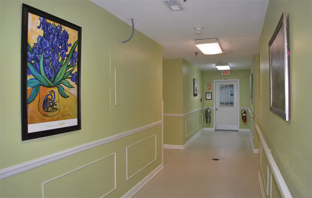 A well-lit pale green interior hallway with framed artwork, handrails, and a door at the far end.