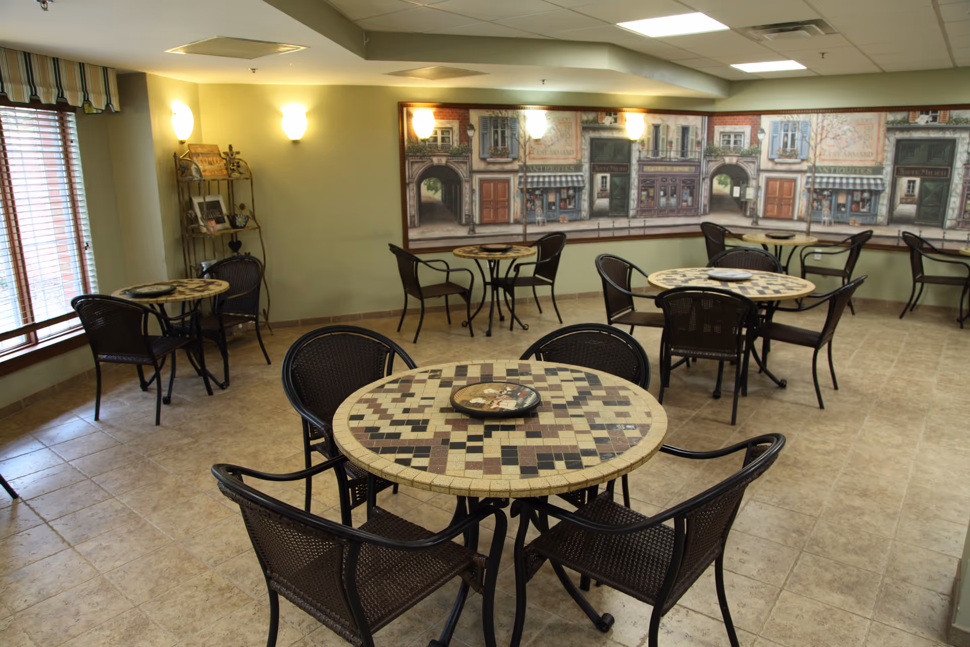 A dining area with several round mosaic-tiled tables and dark wicker chairs arranged around them. The room has tiled floors, light green walls, and a large mural depicting a street scene with storefronts on one wall. There are windows with blinds on the left side and wall-mounted lights providing illumination.