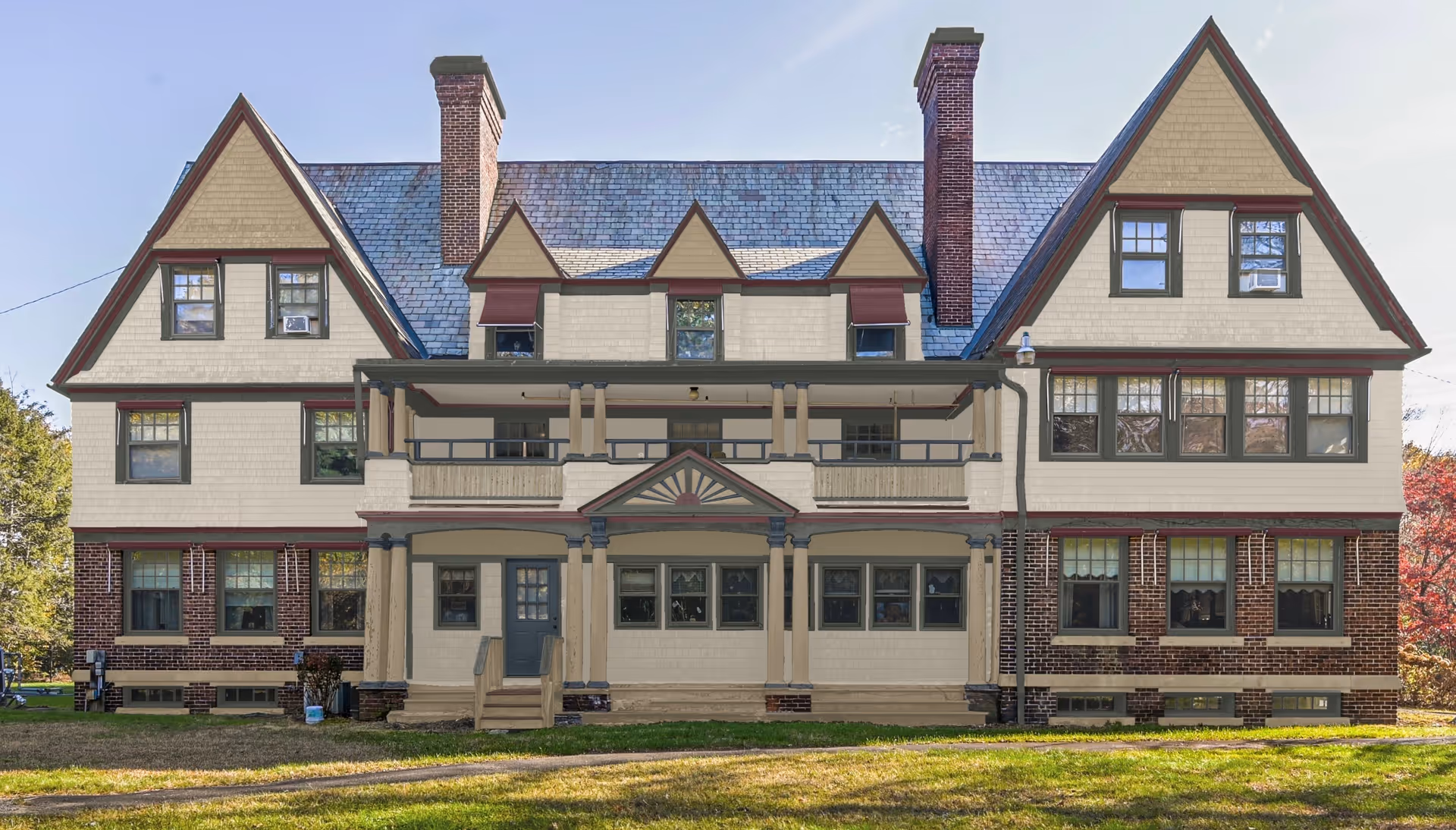 Front exterior view of a large, multi-story assisted living facility building with a steep roof, multiple chimneys, and numerous windows. The building features a combination of brick and light-colored siding with dark trim and a covered porch area.