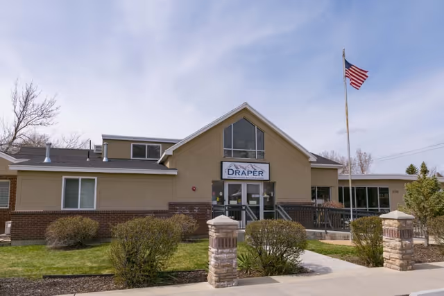 Exterior front view of Draper Rehabilitation and Care Center building with a beige and brick facade, a large window above the entrance, an American flag on a flagpole to the right, and landscaping with bushes and grass in front.