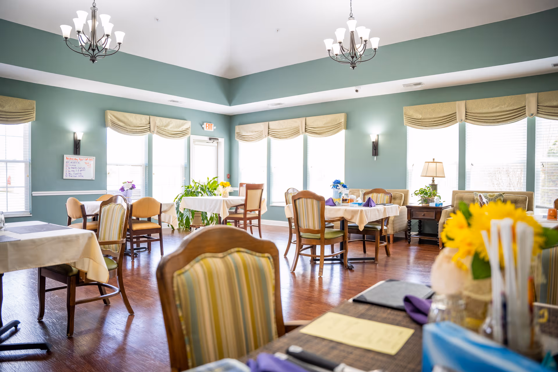 A bright dining room with several tables covered in white tablecloths and set with chairs. The room has large windows with beige valances, green walls, wooden flooring, and two chandeliers hanging from the ceiling. There are plants and flowers on some tables and a side table with a lamp near the windows.