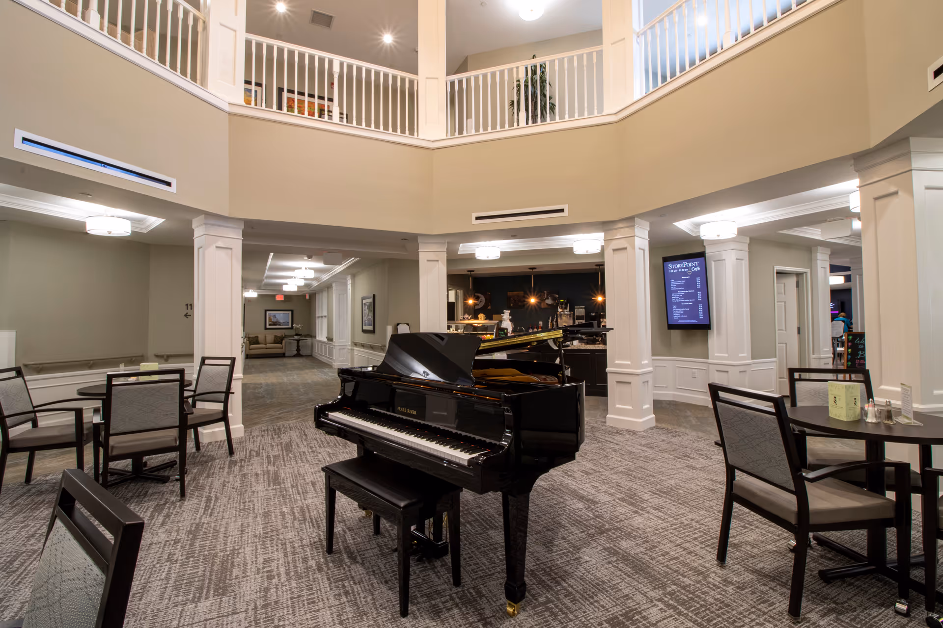 Interior view of a senior living facility common area with a black grand piano in the center, surrounded by tables and chairs. The space has a high ceiling with a second-floor balcony railing visible above. There is a hallway leading to other rooms and a digital menu board on the wall near a café area in the background.