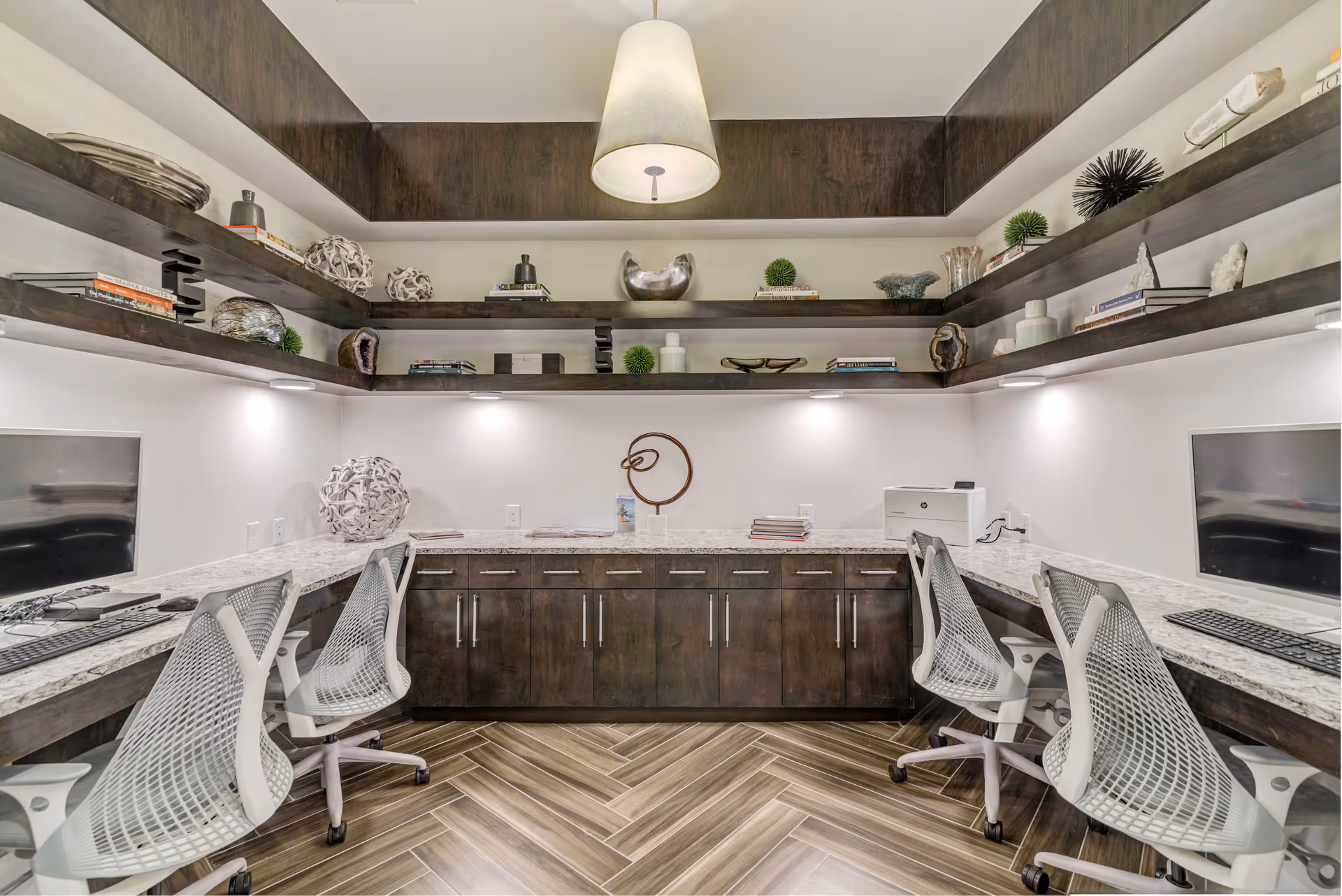 A modern office or computer room with four ergonomic white mesh chairs placed at a U-shaped marble countertop with built-in dark wood cabinets. Two computer monitors and keyboards are positioned on the left and right sides of the countertop. Above the countertop, there are dark wood floating shelves decorated with books, sculptures, and decorative objects. The room has a wood-patterned herringbone floor and a large pendant light hanging from the ceiling.