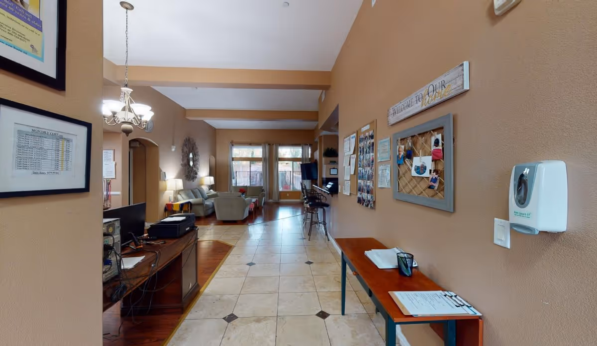 Interior view of a senior living facility hallway with beige walls and tiled floor. On the right wall, there are bulletin boards with photos and notices, a hand sanitizer dispenser, and a wooden table with papers and a cup. On the left side, there is a desk with a computer and office equipment. Further down the hallway, there is a seating area with sofas, lamps, and a large window letting in natural light.