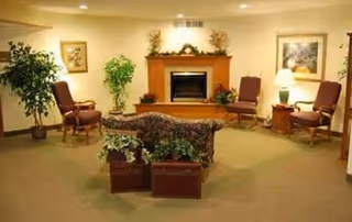 A cozy living room area with a fireplace centered on the back wall, flanked by two framed pictures. There are four upholstered chairs arranged around a small table with a lamp on the right side. Two large potted plants are placed near the walls, and a sofa with a floral pattern faces the fireplace. The room has warm lighting and a carpeted floor.