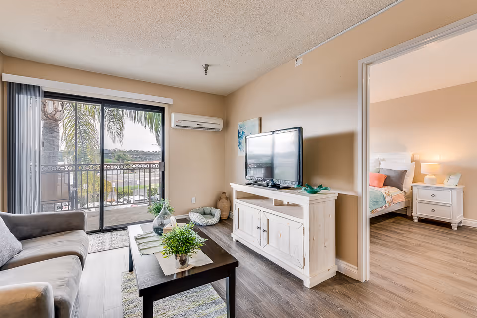 A bright living room with a gray sofa, a dark wooden coffee table with plants on it, and a white TV stand holding a flat-screen television. There is a sliding glass door leading to a balcony with palm trees visible outside. To the right, an open doorway reveals a bedroom with a bed, nightstand, and lamp.