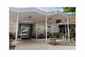 Entrance of a senior living facility named Haven of Meadowbrook with a covered walkway, stone exterior walls, glass doors, potted plants, and outdoor seating including chairs and a table.