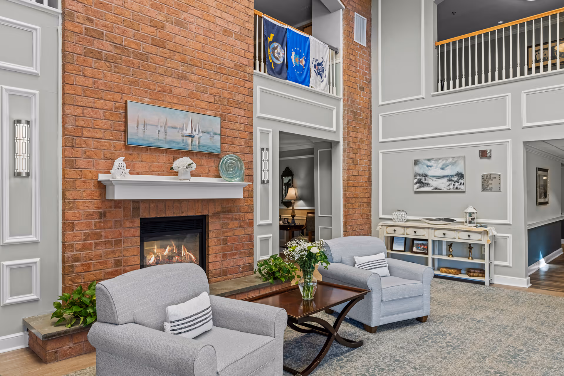A cozy senior living facility common area with two gray armchairs facing a wooden coffee table with a vase of flowers. Behind the chairs is a brick fireplace with a white mantel decorated with a small sculpture, a flower pot, and a decorative plate. Above the mantel is a painting of sailboats on water. The room has high ceilings with a balcony railing above and flags hanging from it. On the right side, there is a white console table with decorative items and framed pictures, and a painting on the wall above it.