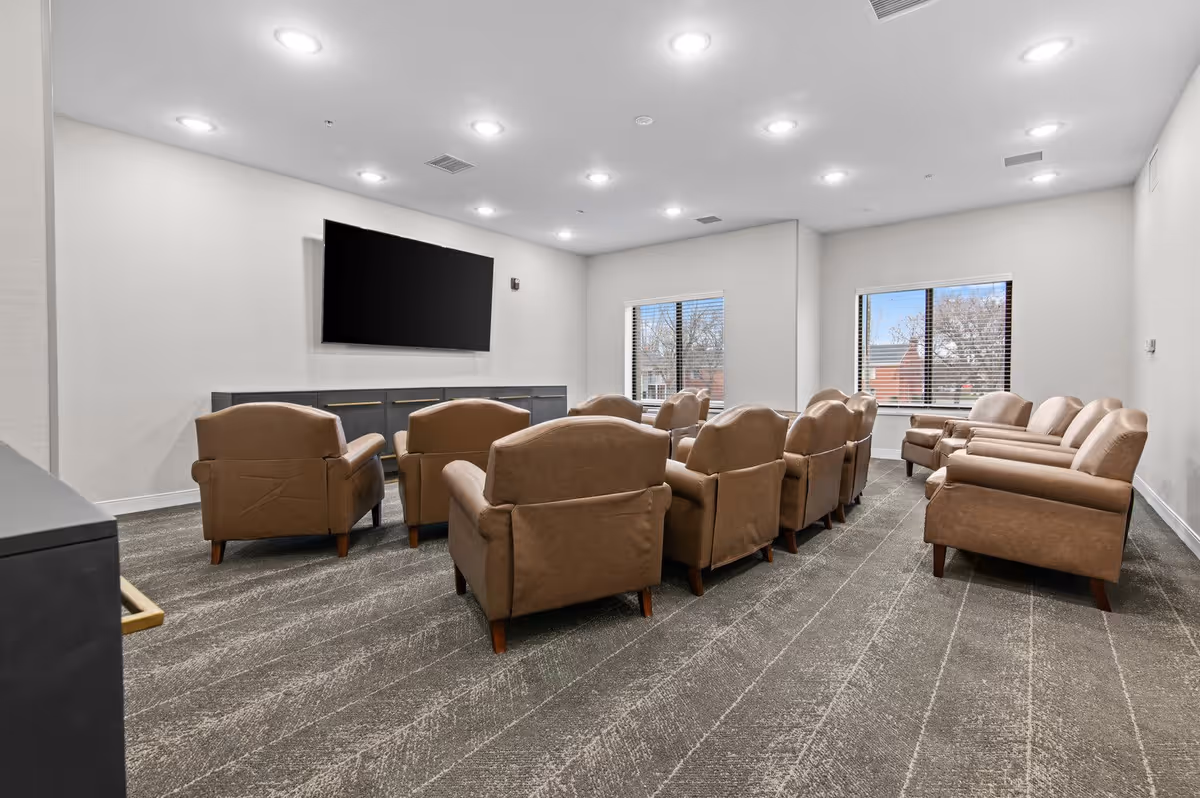 A room with multiple brown leather armchairs arranged in rows facing a large flat-screen TV mounted on a white wall. The room has carpeted flooring, recessed ceiling lights, and two windows with blinds showing an outdoor view.