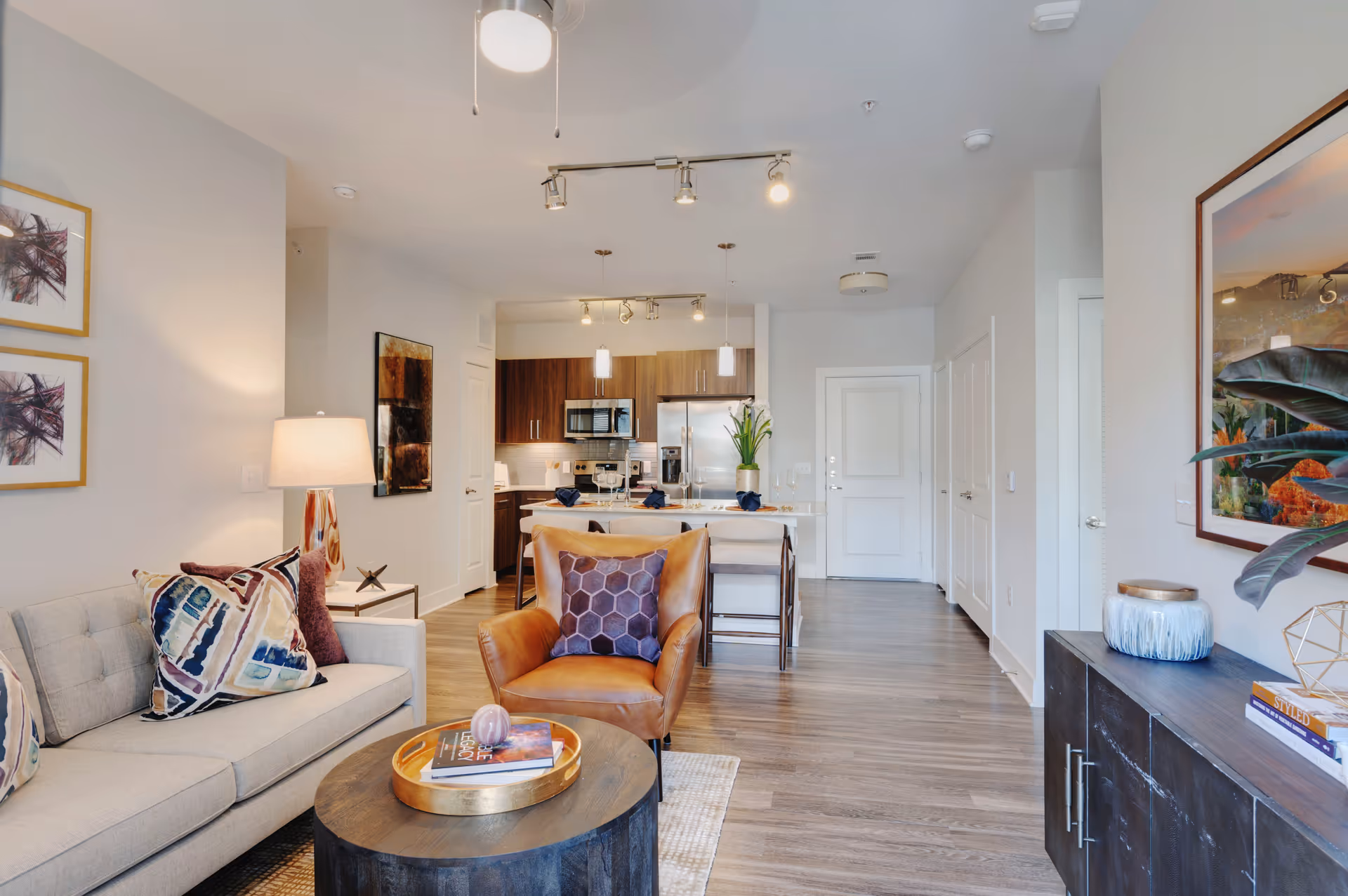 A modern living room and kitchen area with a beige sofa adorned with colorful pillows, a brown leather armchair with a purple cushion, a round wooden coffee table with books and a decorative item, and a kitchen island with bar stools and place settings. The room features wooden flooring, white walls, contemporary lighting fixtures, and framed artwork on the walls.