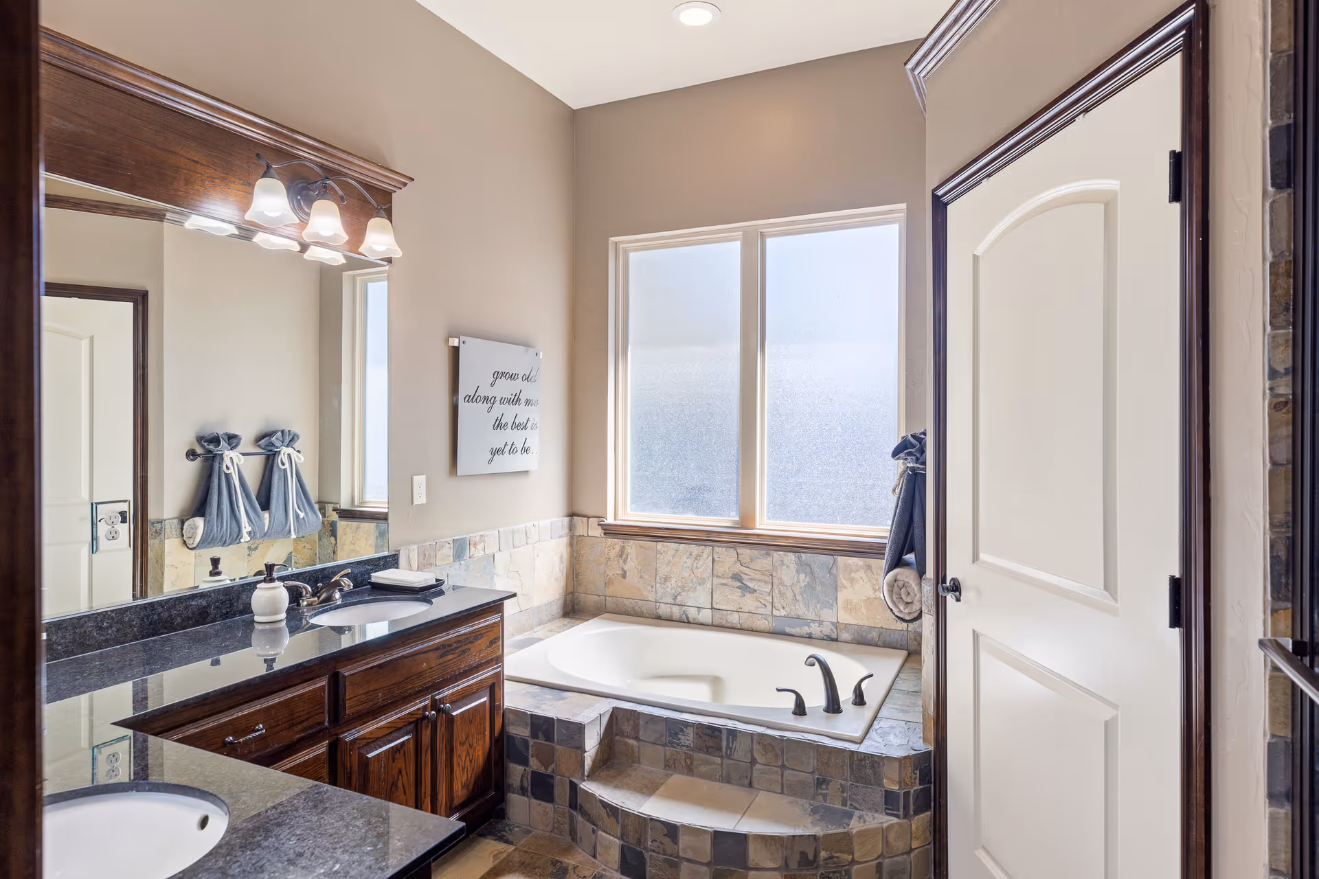 A bathroom featuring a large bathtub with tiled surround and steps, a double sink vanity with dark granite countertops and wooden cabinets, a large mirror with light fixtures above, frosted windows allowing natural light, and a wall decoration with a quote.