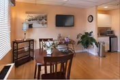 Small dining area with a wooden table and chairs, a wall-mounted TV, decorative plants, and a kitchenette visible in the background.