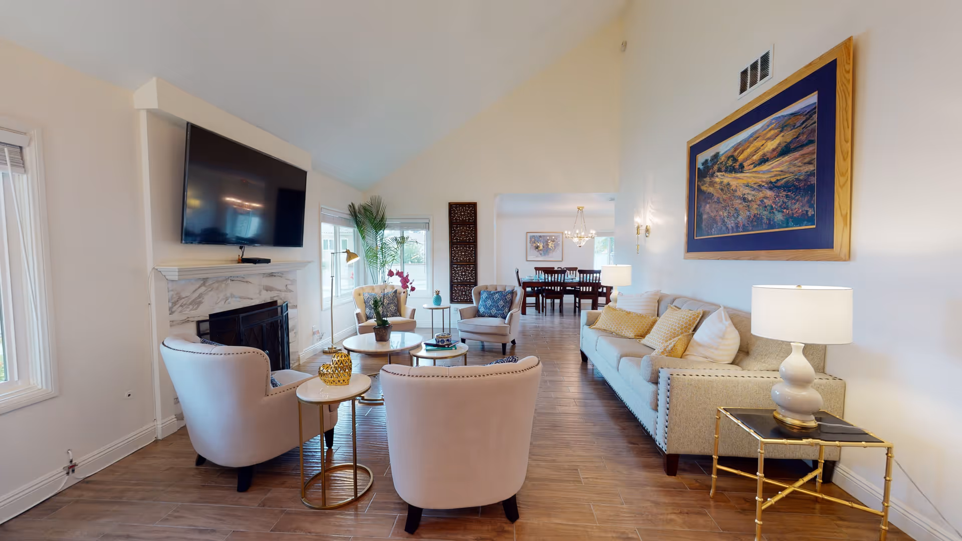 A bright and spacious living room with wooden flooring, featuring a beige sofa with yellow and white cushions, two beige armchairs, a marble fireplace with a mounted flat-screen TV above it, a round coffee table with decorative items, a side table with a lamp, and a large framed landscape painting on the wall. In the background, a dining area with a wooden dining table and chairs is visible under a chandelier.