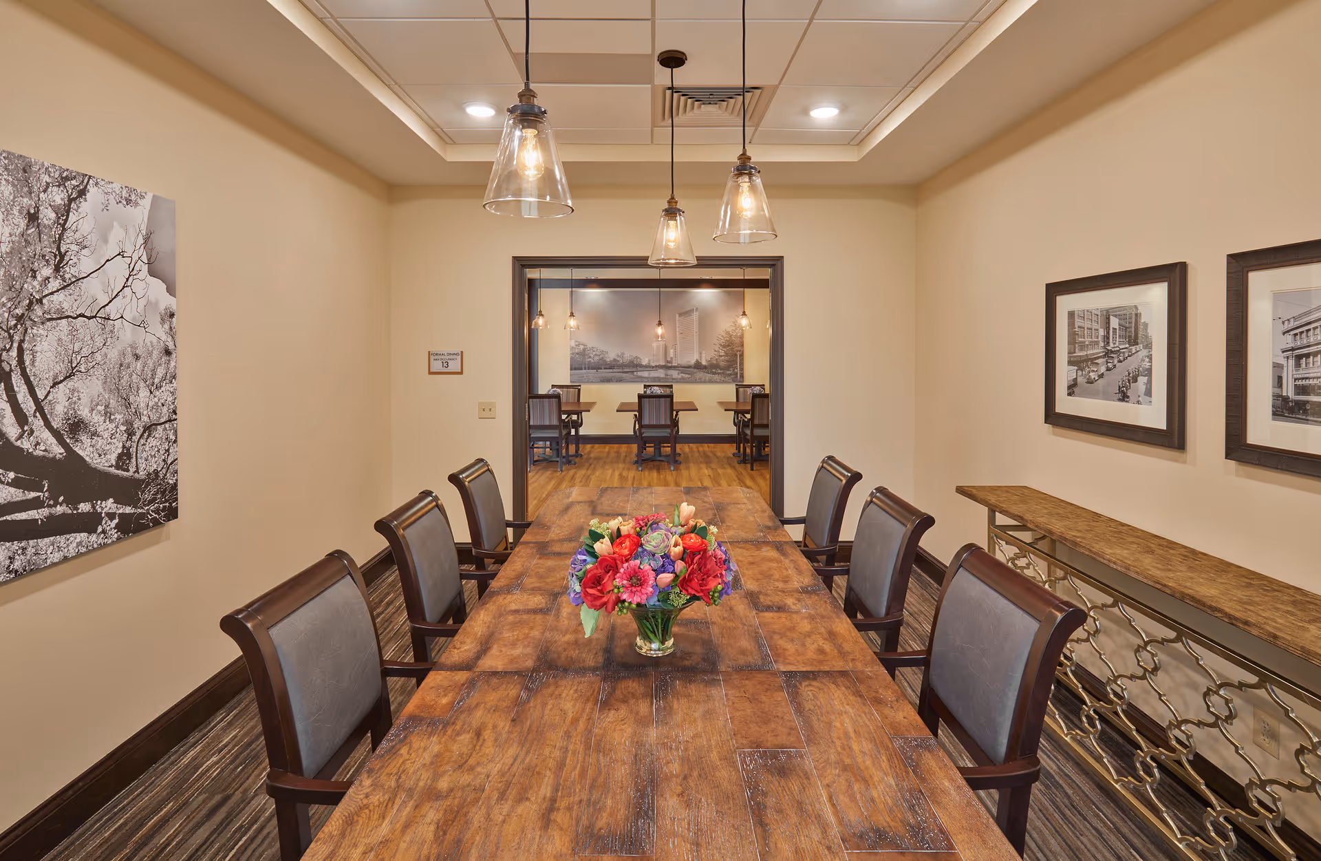 Long wooden dining table with a floral centerpiece surrounded by chairs in a well-lit dining room interior.