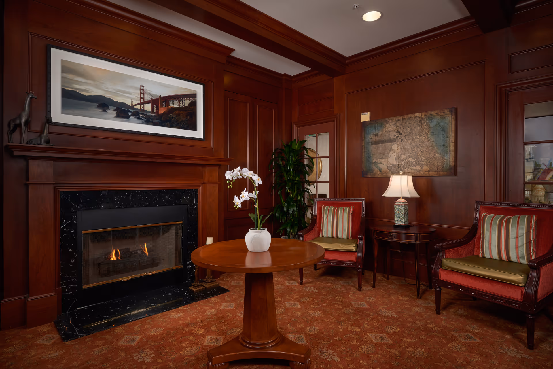 A cozy sitting area with dark wood paneling, a lit fireplace with a black marble surround, and a round wooden table with a white vase holding white orchids. Two red upholstered armchairs with striped cushions flank a small side table with a decorative lamp. A large framed picture of the Golden Gate Bridge hangs above the fireplace, and a vintage map is mounted on the wall behind one of the chairs.