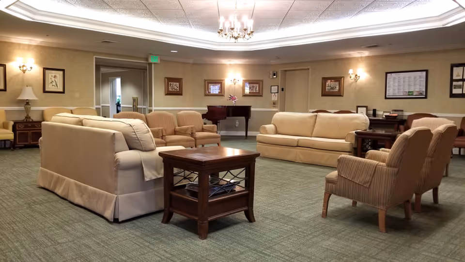 A spacious assisted-living community lounge with beige sofas and armchairs arranged around a wooden coffee table under a chandelier.