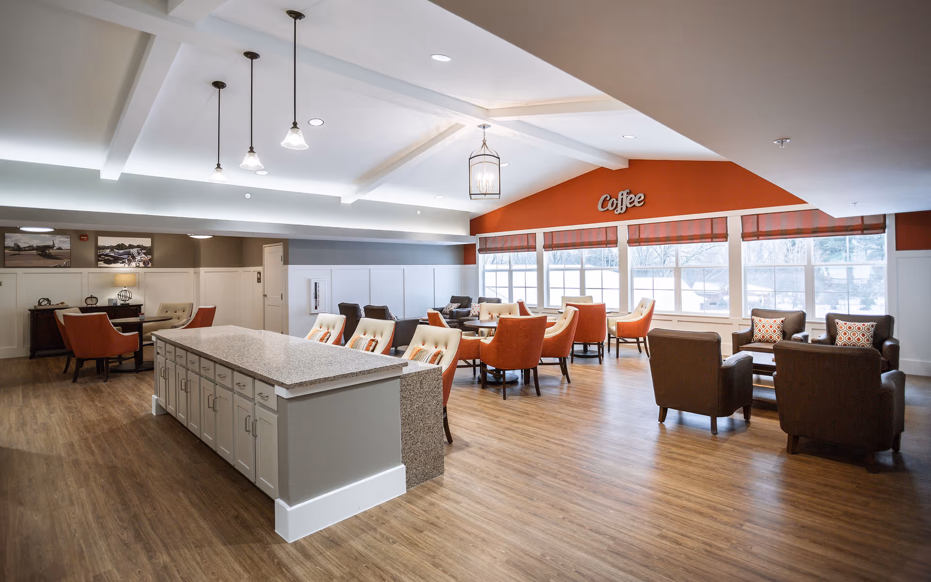 A spacious and well-lit common area in a senior living facility with a large island countertop in the center, surrounded by cushioned chairs. The room features multiple seating arrangements with orange and brown armchairs and small tables. Large windows let in natural light, and the wall above the windows is painted orange with the word 'Coffee' displayed. The floor is wooden, and the ceiling has recessed lighting and hanging pendant lights.