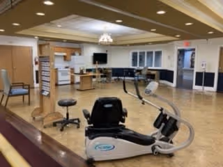 Interior view of a senior living facility common area with exercise equipment including a recumbent bike in the foreground, chairs along the walls, a television mounted on the far wall, and a kitchenette area with cabinets and appliances in the background.