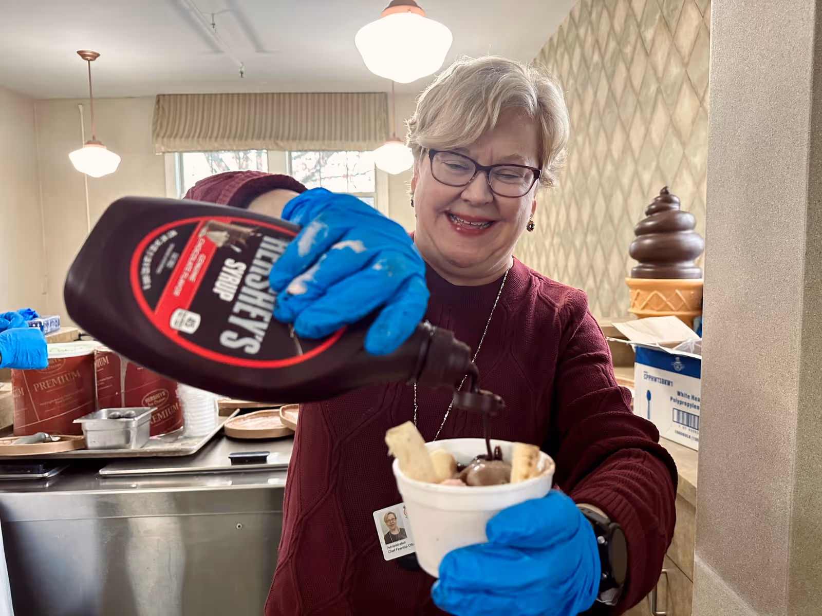 A smiling woman wearing glasses and blue gloves is pouring Hershey's syrup over a cup of ice cream with various toppings in a well-lit room with beige walls and a large ice cream cone decoration in the background.