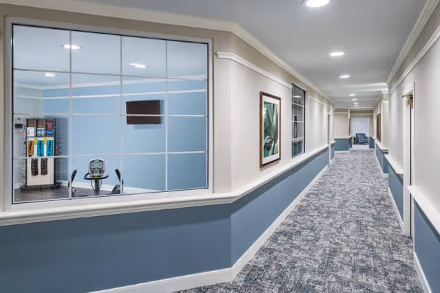 Carpeted interior hallway with blue wainscoting, a large window showing a small fitness room with equipment and a wall-mounted TV, and seating at the far end.