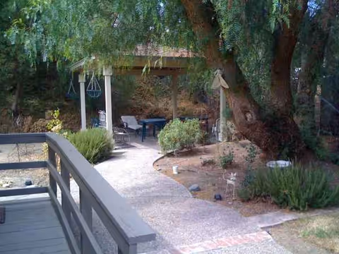 Outdoor courtyard with seating under a shaded gazebo amid trees and shrubs.
