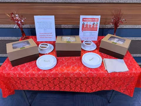 A table covered with a red patterned tablecloth displaying three brown cardboard boxes with clear windows containing food items, three white disposable plates labeled with different food names, two informational signs, two small red vases with decorative red branches, and a stack of white napkins. The table is set against a wall with horizontal wood paneling.
