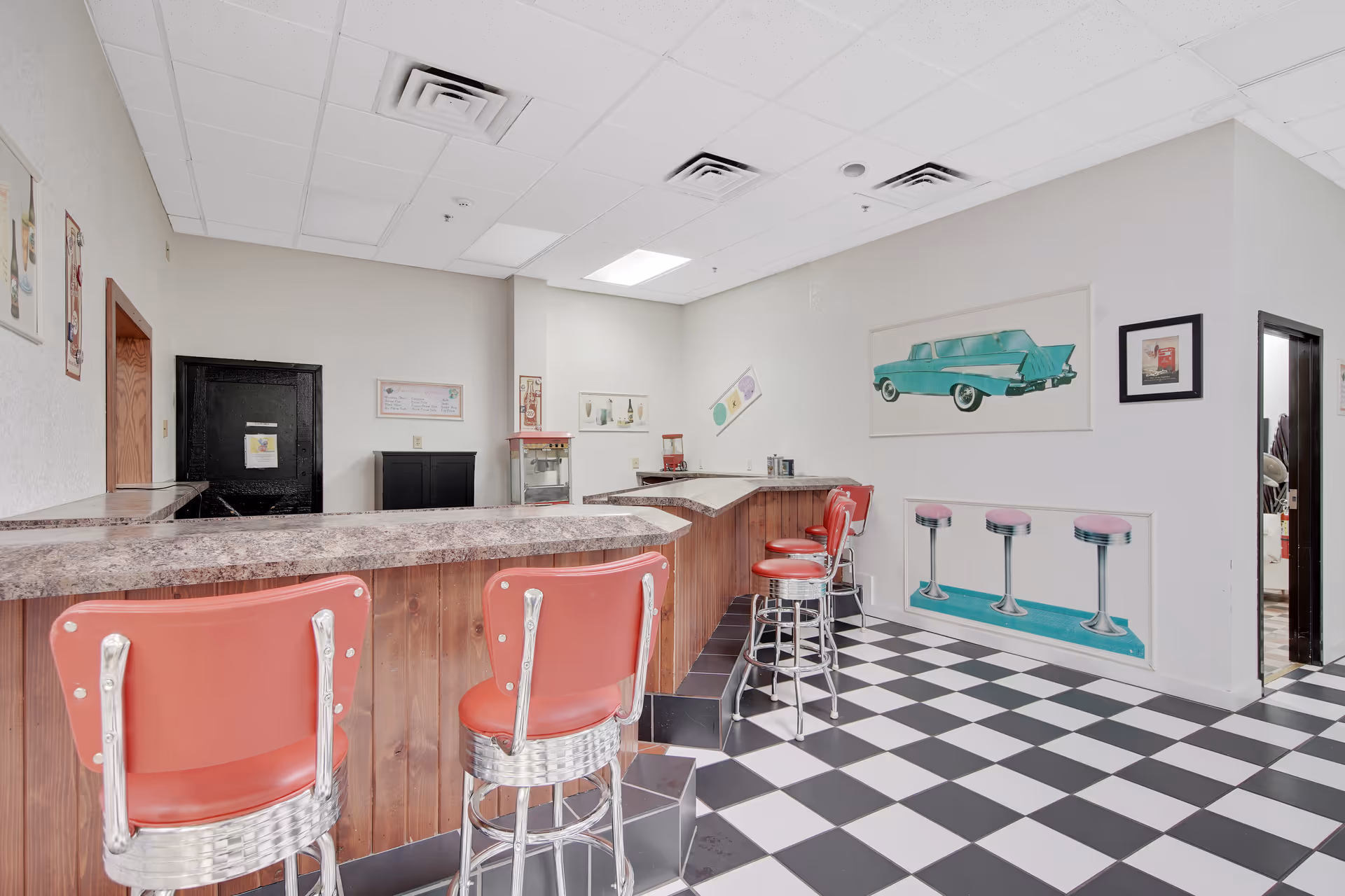 Interior view of a retro-style diner area with a checkered black and white floor, a wooden counter with a marble-like countertop, and red bar stools. The walls are decorated with vintage car and diner-themed artwork. There is a popcorn machine and a soda dispenser on the counter.