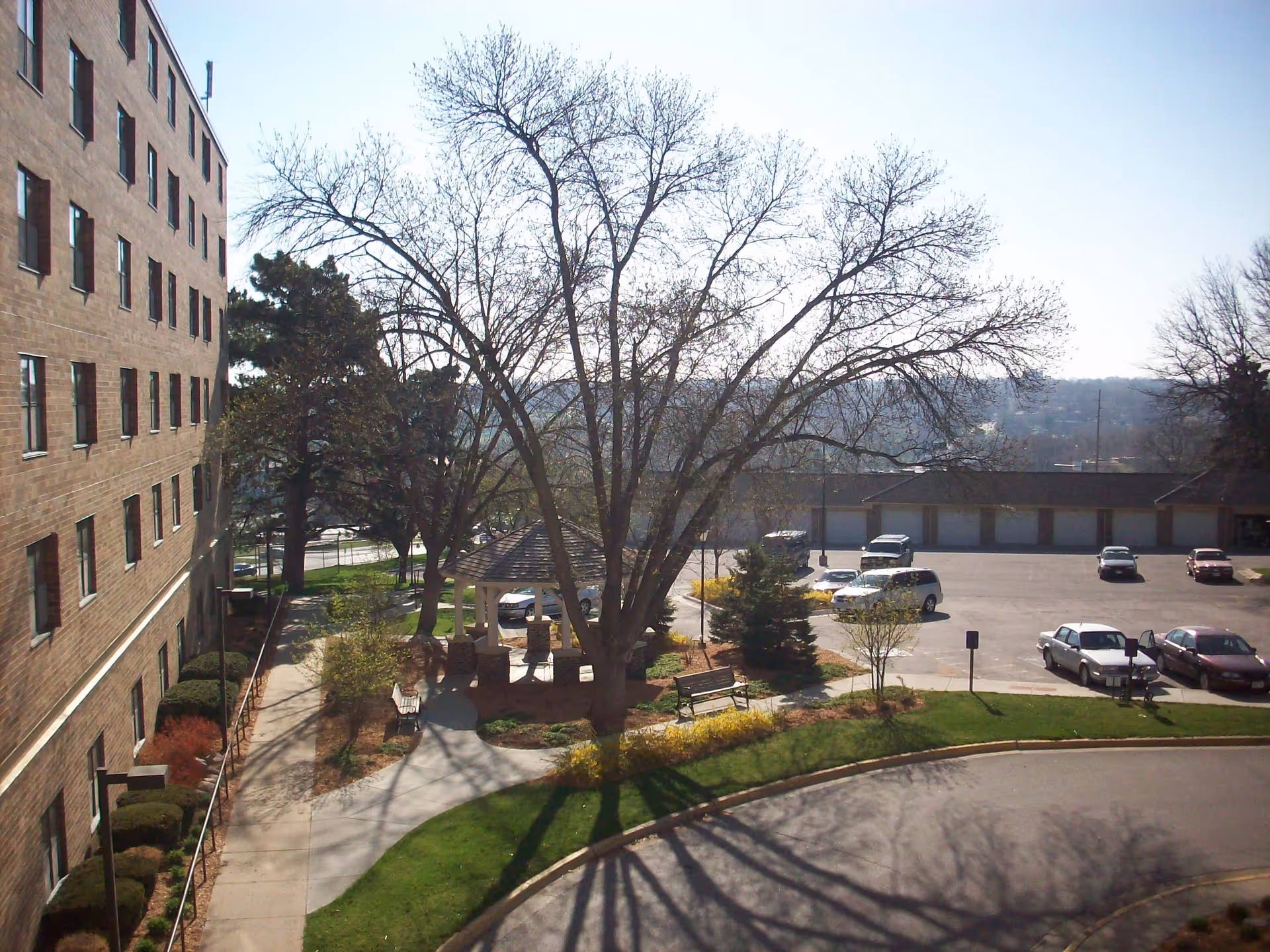 View of an outdoor area at Keystone Villas Senior Living featuring a large tree with bare branches, a gazebo with stone pillars, benches, a curved sidewalk, a parking lot with several cars, and a multi-story brick building on the left side.