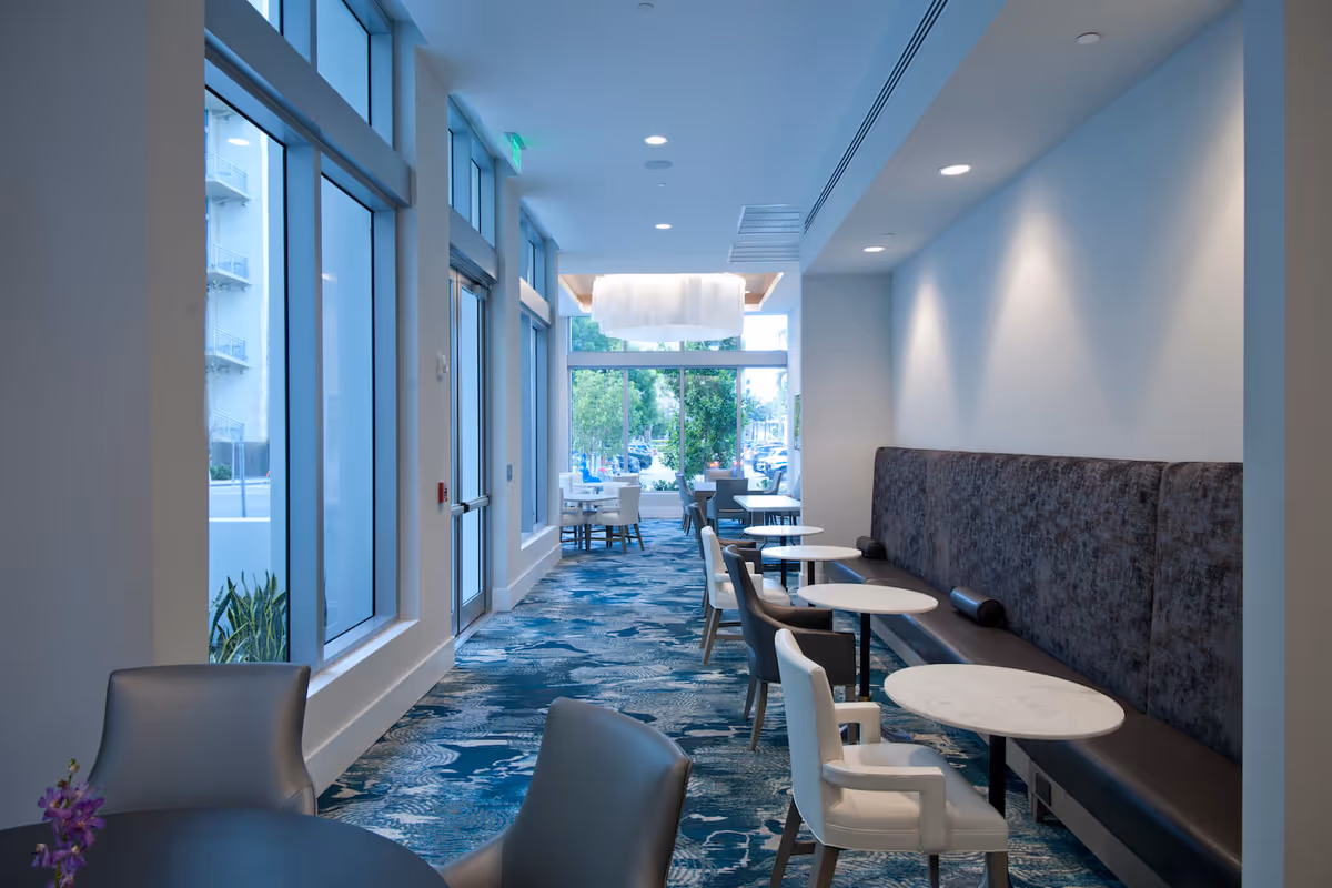 A modern dining area with multiple small round tables and chairs arranged along a wall with a long cushioned bench. Large windows on the left side allow natural light to fill the space, and a decorative ceiling light fixture is visible at the far end near the windows.