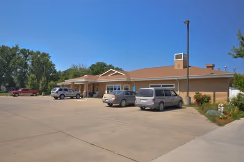 Exterior view of a single-story building with a brown roof and beige siding, surrounded by a large concrete parking lot with several parked cars. There are some trees and shrubs around the building under a clear blue sky.