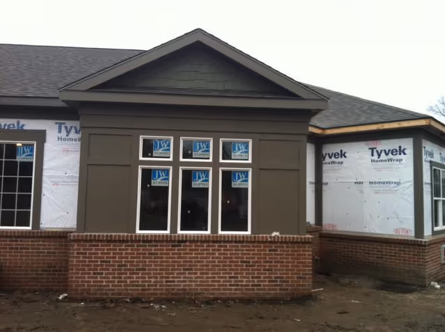 Front exterior of a partially constructed building with a brick knee wall, three windows with manufacturer stickers, and Tyvek-wrapped siding.