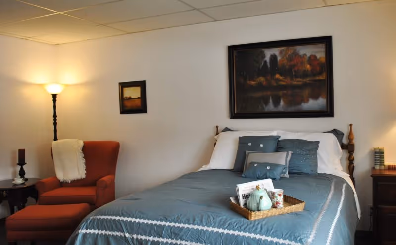 A cozy bedroom with a neatly made bed featuring blue and white bedding and decorative pillows. A tray with a newspaper, a small pumpkin decoration, and a cup sits on the bed. To the left, there is a red armchair with a matching ottoman and a white throw blanket draped over the back. A tall floor lamp and a small side table with a candle and cup are next to the chair. Two framed landscape paintings hang on the white wall above the bed and chair.