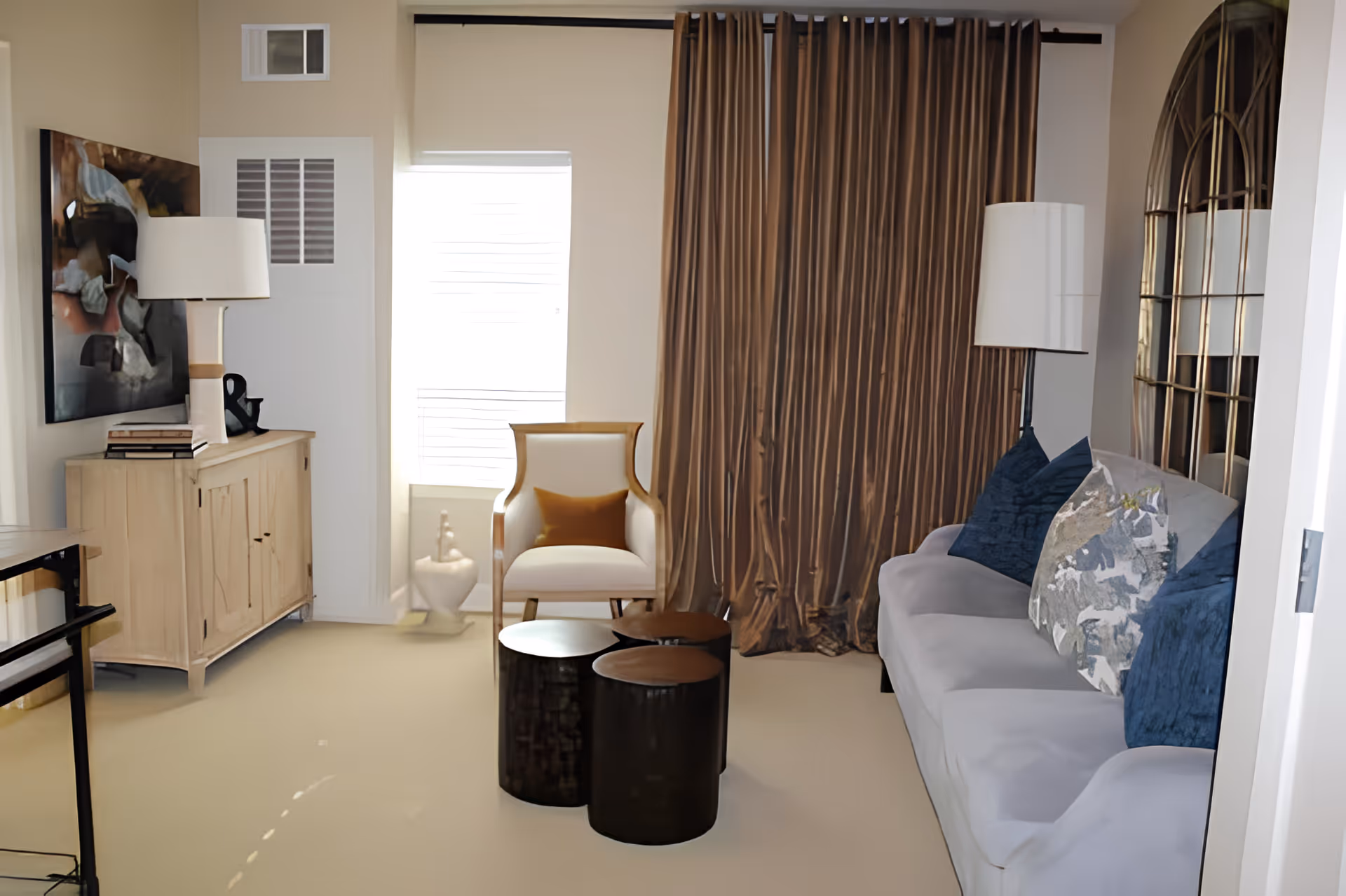 A cozy living room with a light gray sofa adorned with blue and patterned pillows, a white armchair with a brown cushion, three round dark wood coffee tables, a wooden cabinet with a lamp and books, a large decorative mirror on the wall, and brown curtains covering a window.