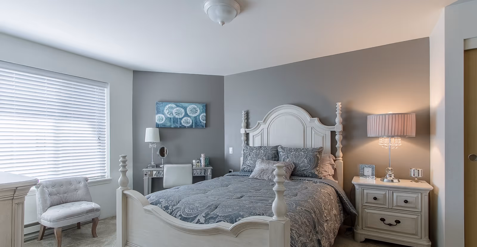 A cozy bedroom with a white wooden bed frame and a gray patterned comforter. There is a nightstand with a lamp and decorative items on the right side of the bed. A small upholstered chair is placed near a window with white blinds on the left side. A small desk with a lamp, mirror, and decorative items is against the gray accent wall behind the bed. The room has soft lighting and a calm, neutral color palette.