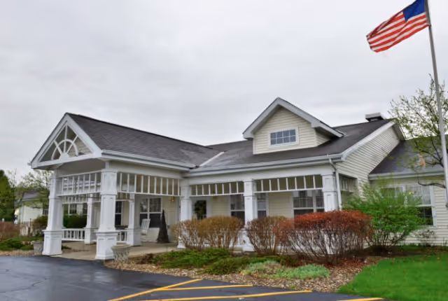 Front entrance of a single-story senior living building with a covered porte-cochere, shrubs, and an American flag.