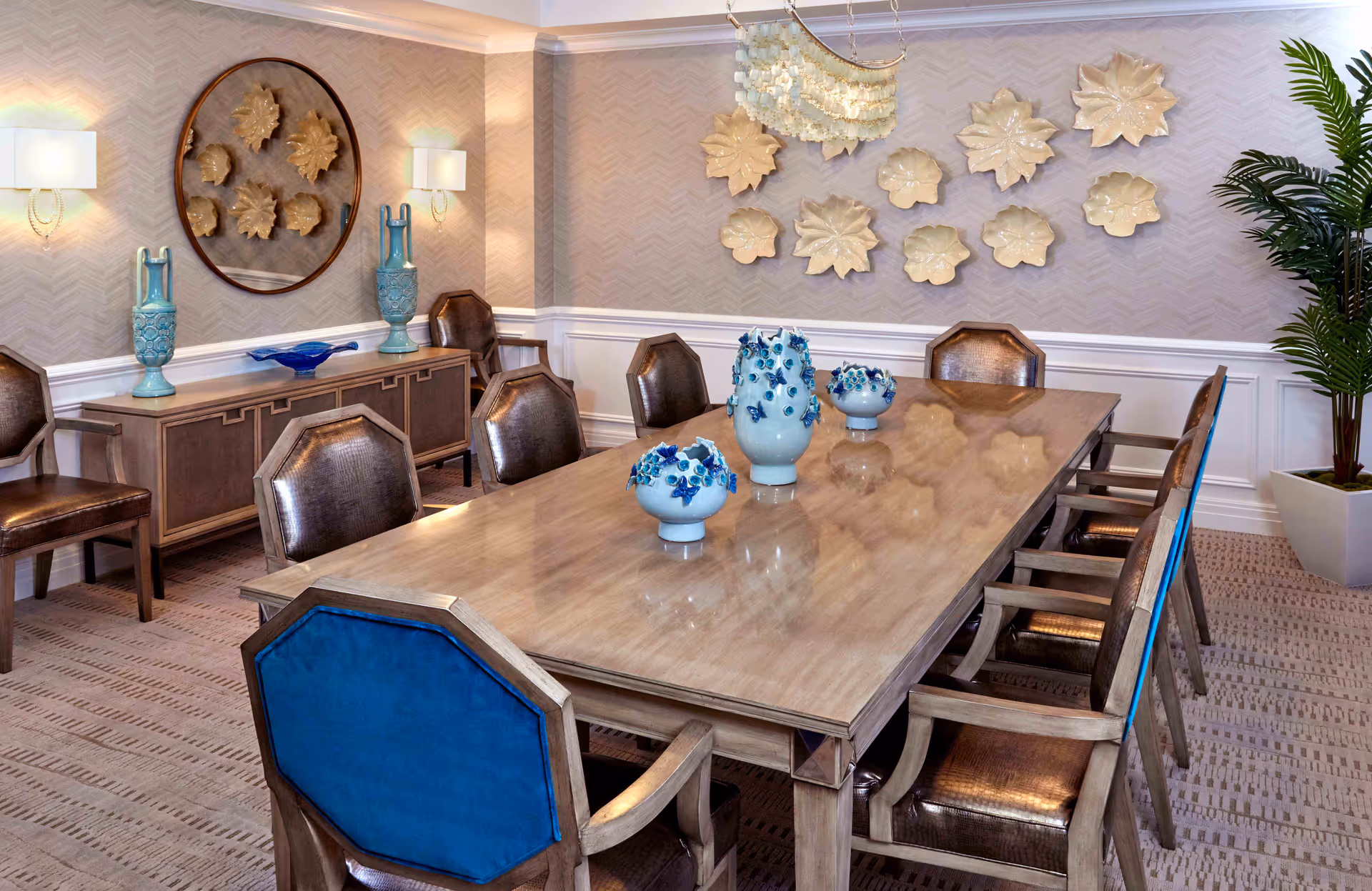 A well-decorated dining room with a long wooden table surrounded by eight chairs, some with blue upholstery on the back. The table has three decorative blue and white vases. The walls feature beige patterned wallpaper with white wainscoting and are adorned with decorative ceramic wall art and a round mirror. Two wall sconces with lampshades provide lighting, and a large potted plant is in the corner.