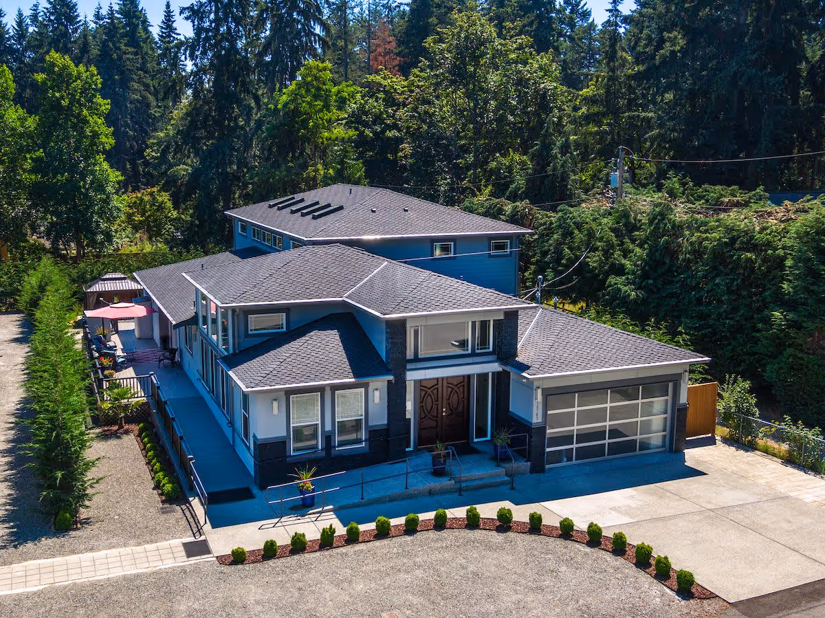 Aerial view of a modern two-story residential building with a garage, driveway, front entrance, and surrounding trees.