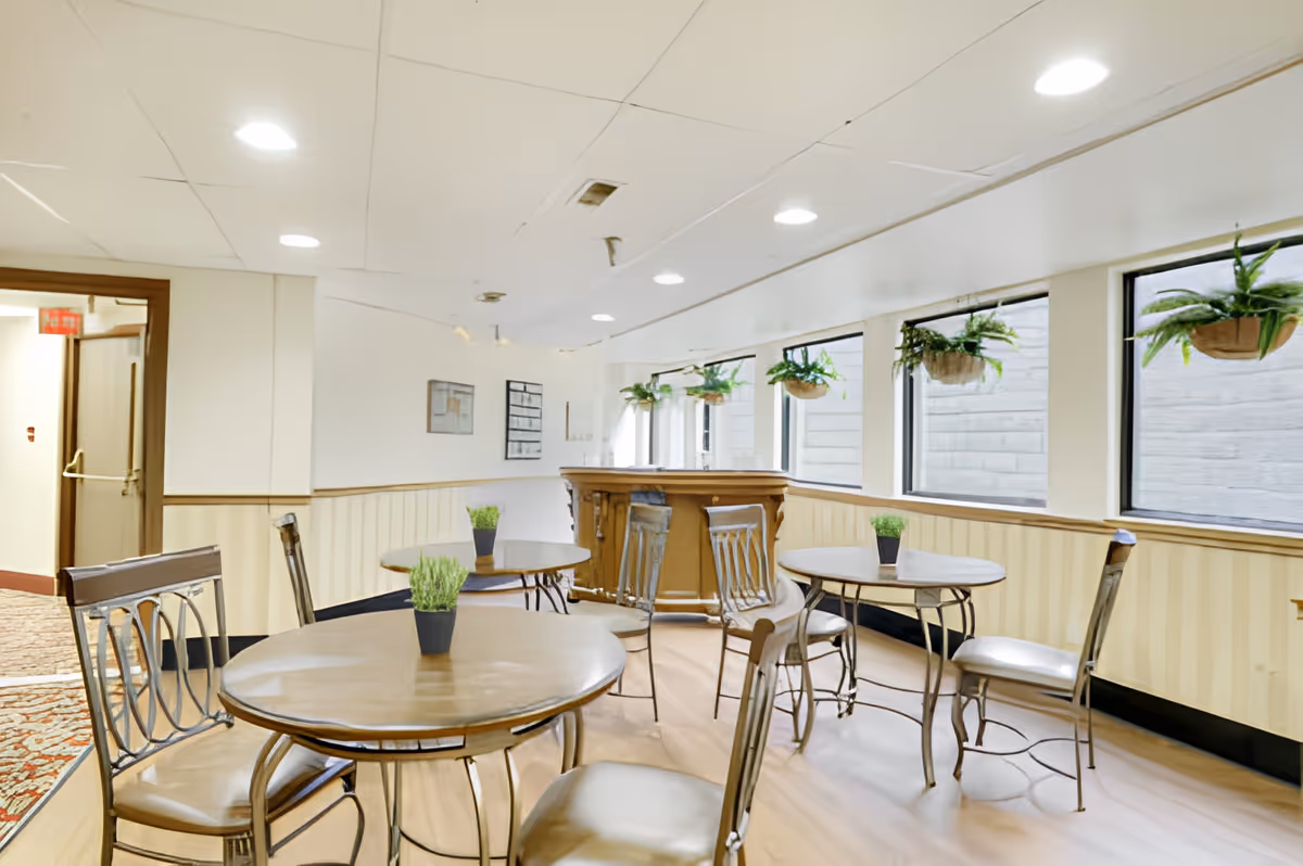 Small communal dining area with round tables and chairs, potted plants, and windows along one wall.