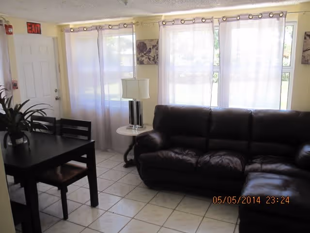 A living room area with a dark brown leather sectional sofa, a small round side table with a lamp, and a black dining table with chairs. The room has tiled floors and large windows covered with sheer white curtains, allowing natural light to enter. There is an exit door on the left side of the room.