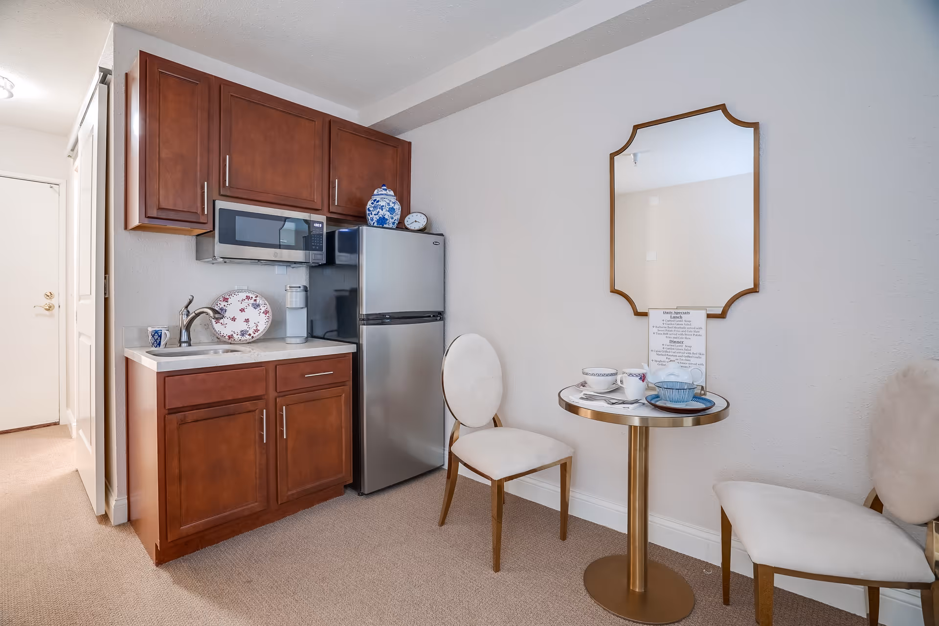 Small kitchenette with wooden cabinets, a microwave and mini fridge beside a small round table and two chairs under a wall mirror.