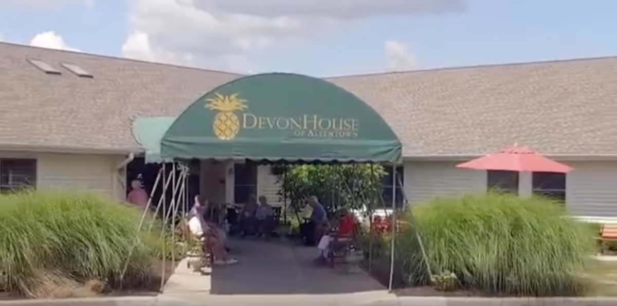 Entrance to DevonHouse Senior Living Allentown with a green canopy featuring a pineapple logo and the facility name. Several seniors are seated outside under the canopy and near the building, surrounded by tall grass and a pink umbrella on the right side.
