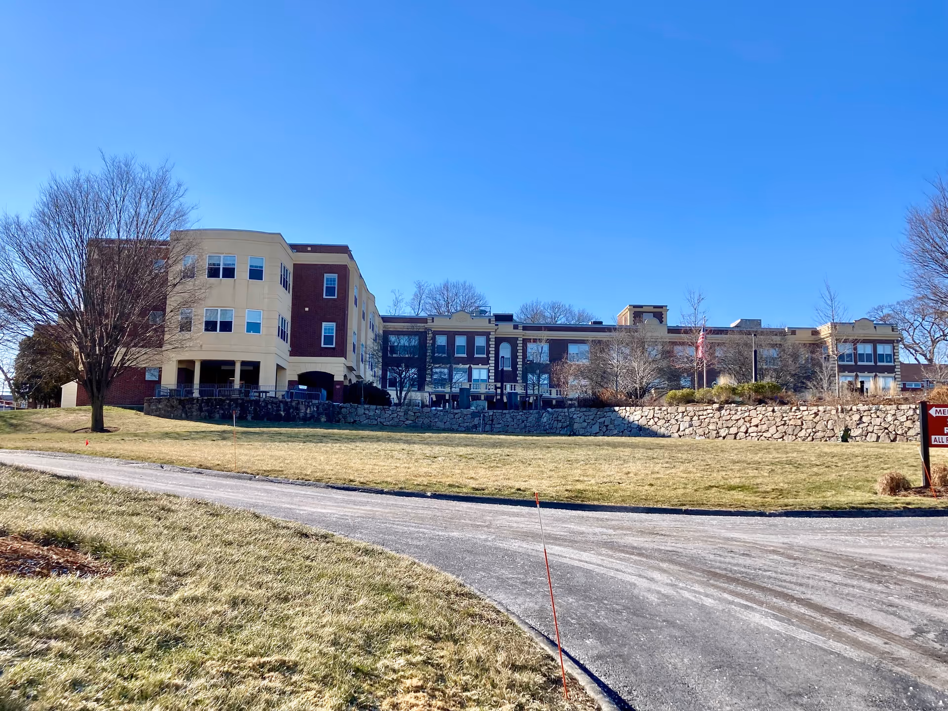 Exterior view of a multi-story senior living facility building with a mix of brick and beige siding, surrounded by leafless trees and a grassy area under a clear blue sky.