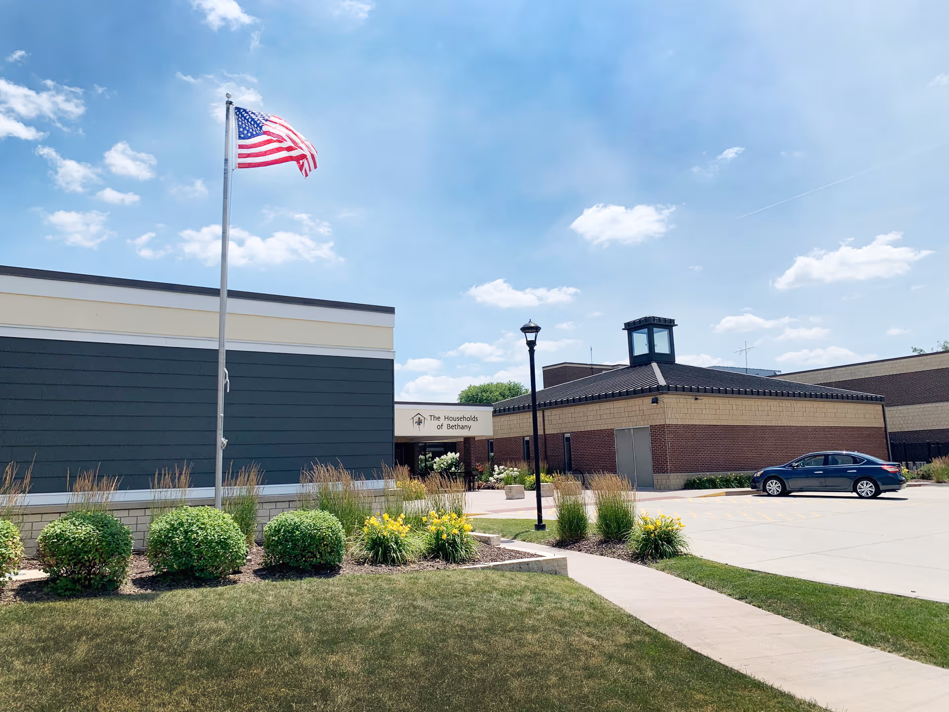 Exterior front of a Bethany Life senior living building with an American flag, landscaped lawn, and a parked car.