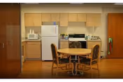 A small kitchen area with light wood cabinets, a white refrigerator, microwave, stove, and oven. In front of the kitchen is a round wooden table with four cushioned chairs. The floor is wooden, and there is a fire extinguisher mounted on the wall to the right.