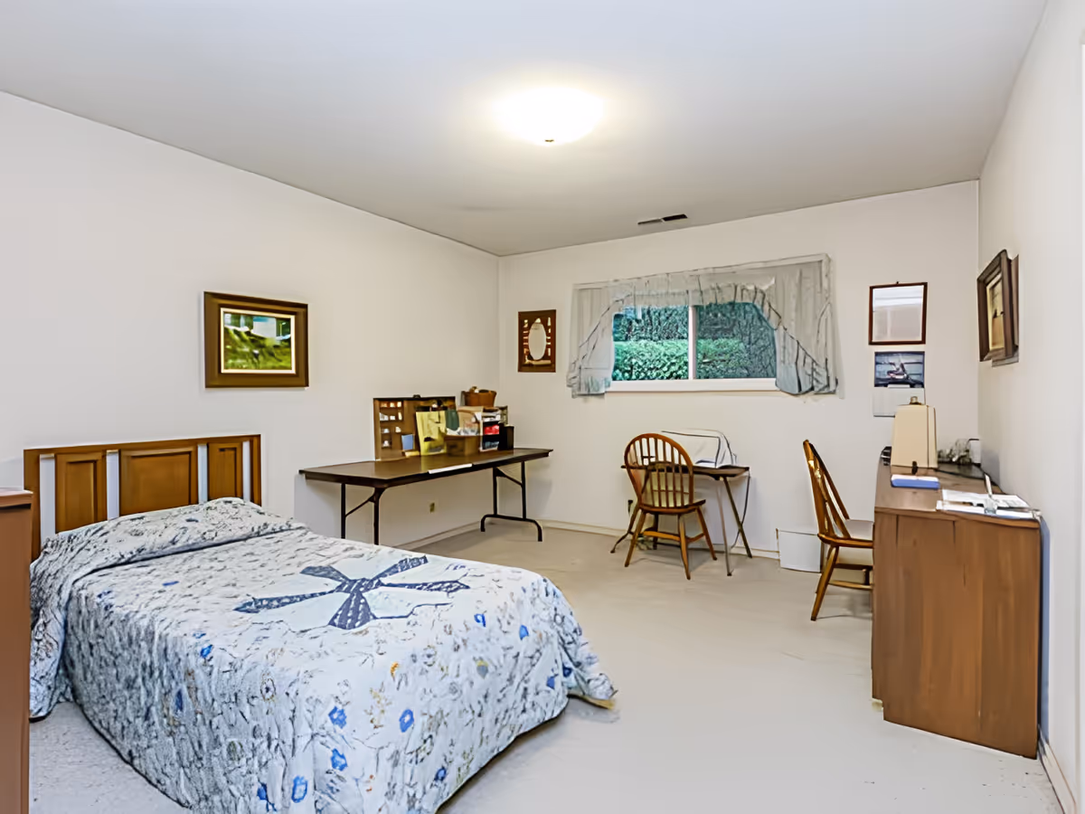 A simple bedroom with a single bed covered in a patterned quilt, a long table with various items on it, two wooden chairs, a small desk, a dresser with a lamp, and a window with curtains showing greenery outside. The walls are white and decorated with a few framed pictures.